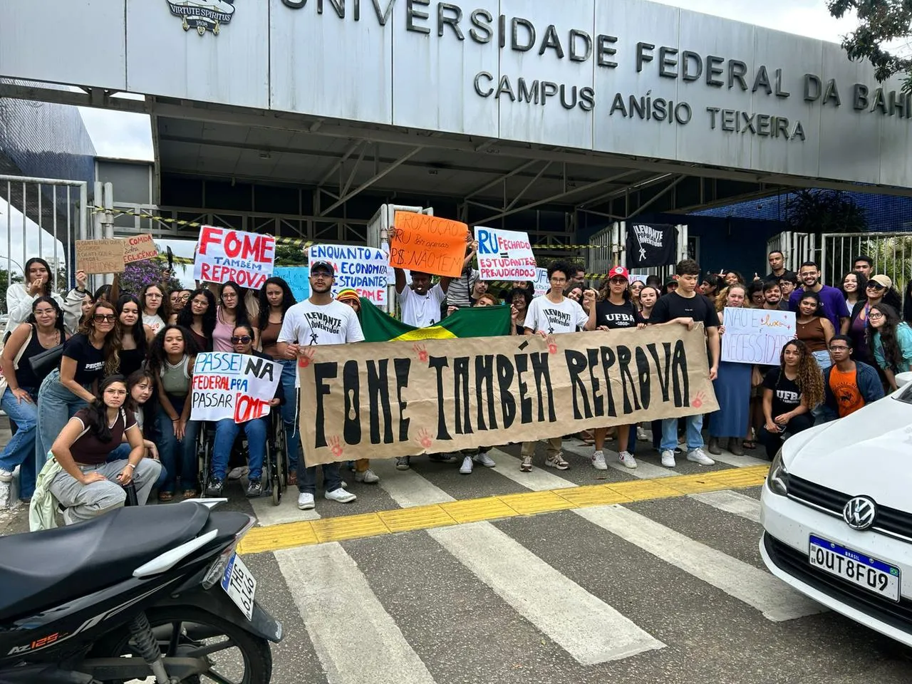Grupo de estudantes protesta em frente à entrada da Universidade Federal da Bahia, Campus Anísio Teixeira. No centro, várias pessoas seguram uma faixa bege com a frase “FOME TAMBÉM REPROVA”. Outros cartazes dizem: “Fome também reprova”, “RU fechado = estudantes prejudicados”, “Passei na Federal para passar fome”, “Promoção RU dos sonhos R$ não tem” e “Enquanto a economia, estudantes com fome”. Há uma bandeira do Brasil atrás da faixa principal. Os estudantes usam roupas casuais e estão reunidos sobre uma faixa de pedestres. Ao fundo aparece a fachada da universidade.