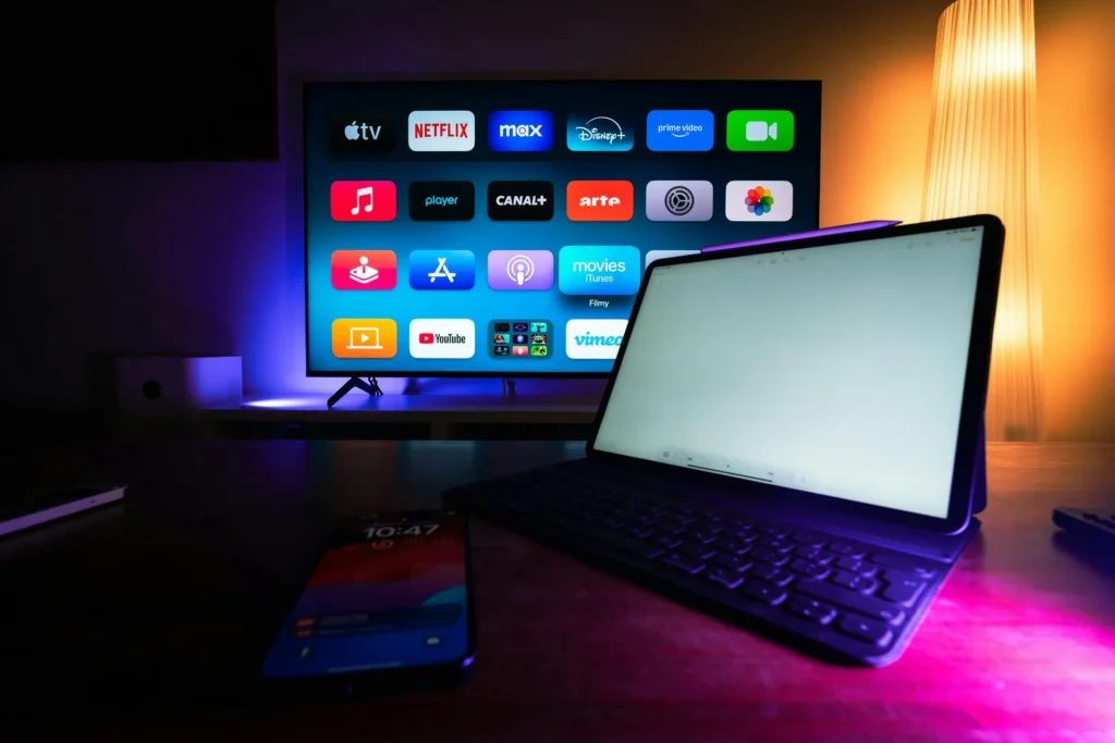 Em um ambiente escuro iluminado por luzes em tons de azul, rosa e amarelo, uma televisão exibe a tela inicial aplicativos da Netflix, Disney+, Prime Video e YouTube. À frente da TV, sobre uma mesa de madeira, há um tablet com teclado acoplado e a tela clara ligada. Um celular aparece desfocado no primeiro plano. À direita, uma luminária alta emite luz amarelada. Fonte: Jakub Zerdzicki.