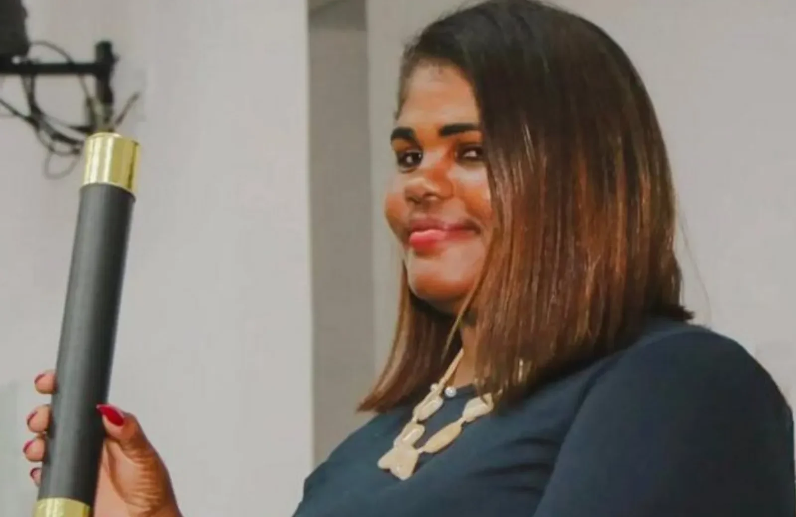 Mulher negra sorridente segura um canudo de formatura preto com extremidades douradas. Ela tem cabelos castanhos lisos na altura dos ombros, usa unhas vermelhas, um colar e blusa azul-marinho.