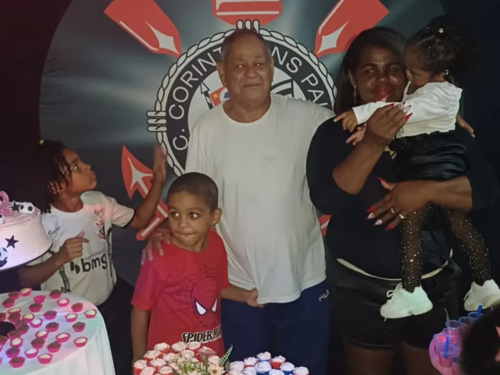 Um homem e uma mulher posam sorridentes ao centro de uma festa de aniversário com três crianças. O homem veste camiseta branca e a mulher usa blusa preta, segurando uma criança pequena no colo. À esquerda, um menino usa camiseta do Corinthians e outro veste camiseta vermelha do Homem-Aranha. À frente deles, há mesas com um bolo branco decorado com estrelas, docinhos e flores.