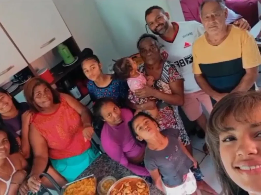 Grupo de pessoas negras de diversas idades posa para uma foto em uma cozinha. No centro, uma mulher segura uma criança pequena no colo, cercada por homens, mulheres e outras crianças sorridentes. À frente deles, sobre uma mesa de granito escuro, há travessas com comida.