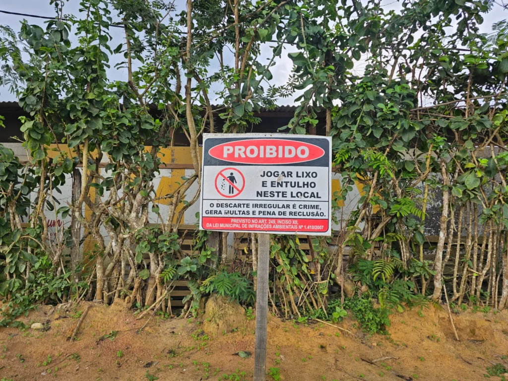 Uma placa de sinalização retangular está fixada sobre um chão de terra batida. A placa possui fundo branco, bordas pretas e traz um símbolo de proibição com uma silhueta humana descartando lixo riscada em vermelho. O texto impresso na placa diz: "PROIBIDO JOGAR LIXO E ENTULHO NESTE LOCAL O DESCARTE IRREGULAR É CRIME, GERA MULTAS E PENA DE RECLUSÃO. PREVISTO NO ART. 249, INCISO XII, DA LEI MUNICIPAL DE INFRAÇÕES AMBIENTAIS Nº 1.410/2007".