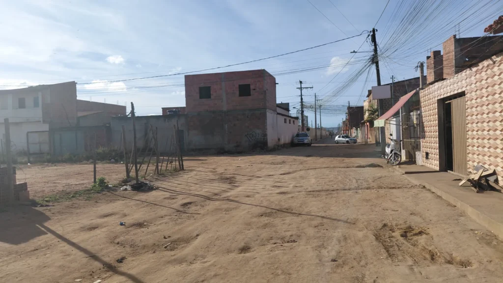 Uma rua de terra larga se estende em direção a uma casa de dois andares com paredes de tijolos vermelhos. No lado direito, há casas com fachadas rebocadas, uma motocicleta branca estacionada na calçada, postes de iluminação e um grande emaranhado de fios elétricos no céu. Ao fundo da rua, dois carros estão estacionados. 