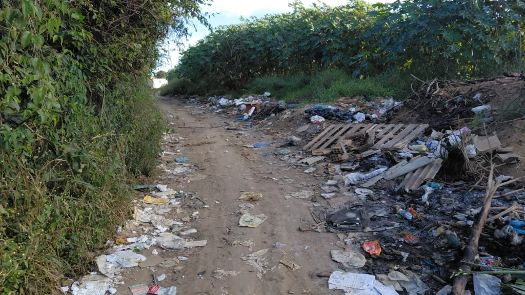 Uma estrada de terra é cercada por uma grande quantidade de lixo e entulho. Os resíduos incluem sacolas plásticas, papéis, embalagens coloridas, restos de madeira e um palete de madeira velho. Paredes densas de vegetação verde e arbustos altos cercam os dois lados da via.
