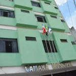 Fachada da Câmara Municipal de Vitória da Conquista vista de baixo para cima. O prédio possui vários andares com paredes na cor verde-claro, detalhes em branco e janelas de vidro escuro. Na parte central, três bandeiras estão hasteadas em mastros verticais: a da Bahia, a do Brasil e a de Vitória da Conquista.