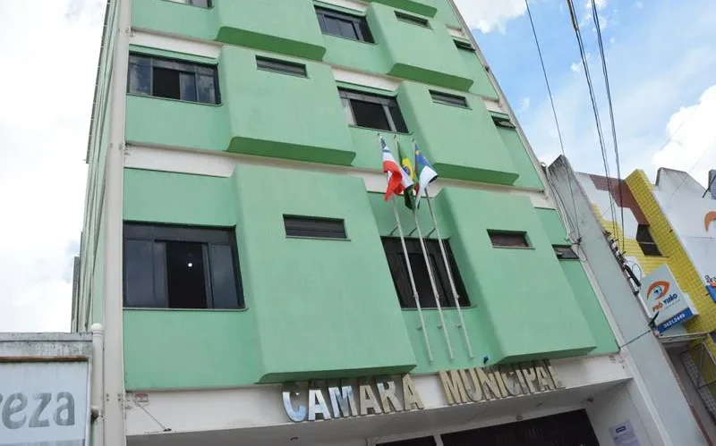Fachada da Câmara Municipal de Vitória da Conquista vista de baixo para cima. O prédio possui vários andares com paredes na cor verde-claro, detalhes em branco e janelas de vidro escuro. Na parte central, três bandeiras estão hasteadas em mastros verticais: a da Bahia, a do Brasil e a de Vitória da Conquista.
