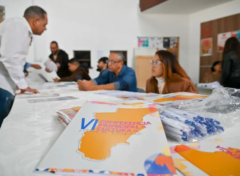 Uma brochura está em primeiro plano sobre uma mesa. O texto nela diz: "6ª CONFERÊNCIA MUNICIPAL DE CULTURA". Ao lado há canetas em um saco plástico. Ao fundo, pessoas sentam-se à mesa e um homem de camisa branca está de pé. Paredes brancas com pôsteres formam o cenário.
