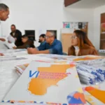 Uma brochura está em primeiro plano sobre uma mesa. O texto nela diz: "6ª CONFERÊNCIA MUNICIPAL DE CULTURA". Ao lado há canetas em um saco plástico. Ao fundo, pessoas sentam-se à mesa e um homem de camisa branca está de pé. Paredes brancas com pôsteres formam o cenário.