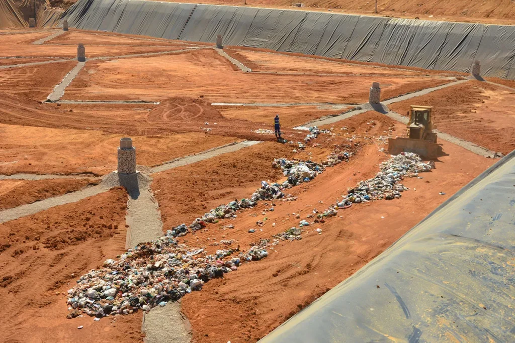 Um trator empurra uma trilha de lixo doméstico sobre o solo de terra vermelha de um aterro sanitário. Ao fundo e nas laterais direita e superior, grandes encostas escavadas estão completamente cobertas por uma lona plástica.