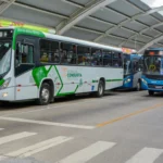 Dois ônibus urbanos estão estacionados em um terminal coberto com estrutura metálica.