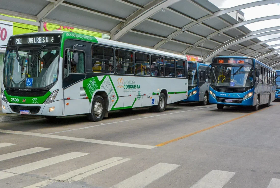 Dois ônibus urbanos estão estacionados em um terminal coberto com estrutura metálica.