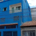 Prédio residencial de três andares pintado em azul. O edifício possui janelas com grades no térreo e no primeiro andar. No segundo andar, há uma sacada recuada com parede interna branca, porta e janela verde-claras protegidas por uma grade de ferro.