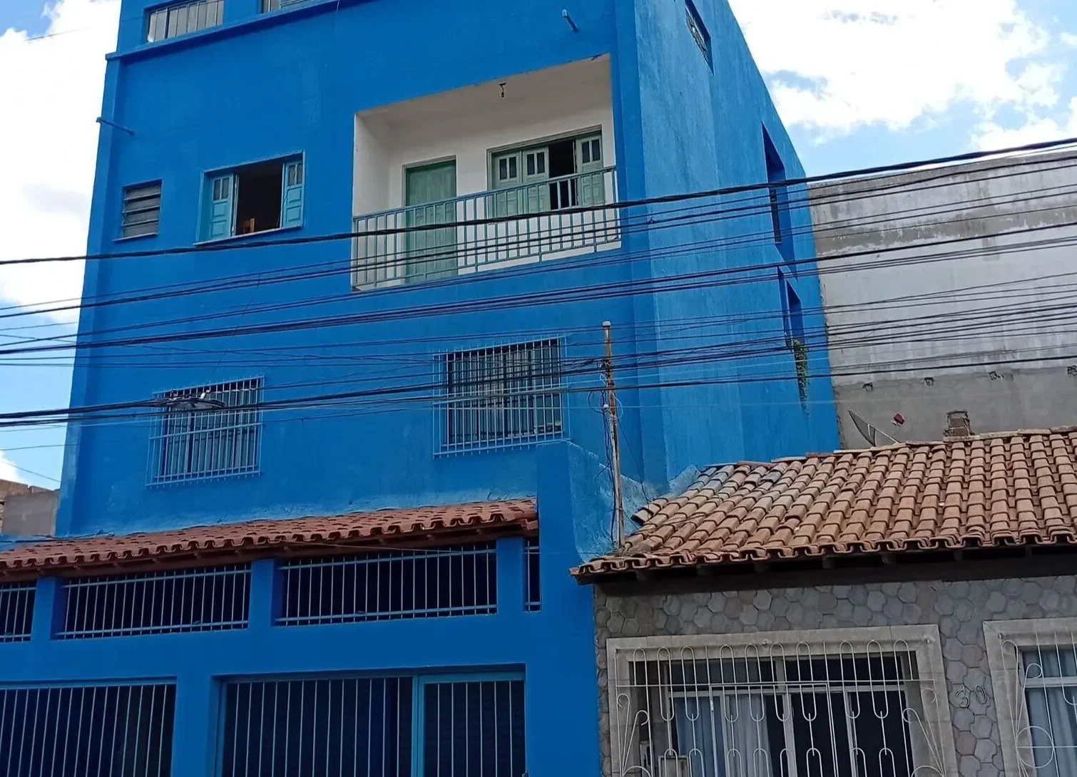 Prédio residencial de três andares pintado em azul. O edifício possui janelas com grades no térreo e no primeiro andar. No segundo andar, há uma sacada recuada com parede interna branca, porta e janela verde-claras protegidas por uma grade de ferro.
