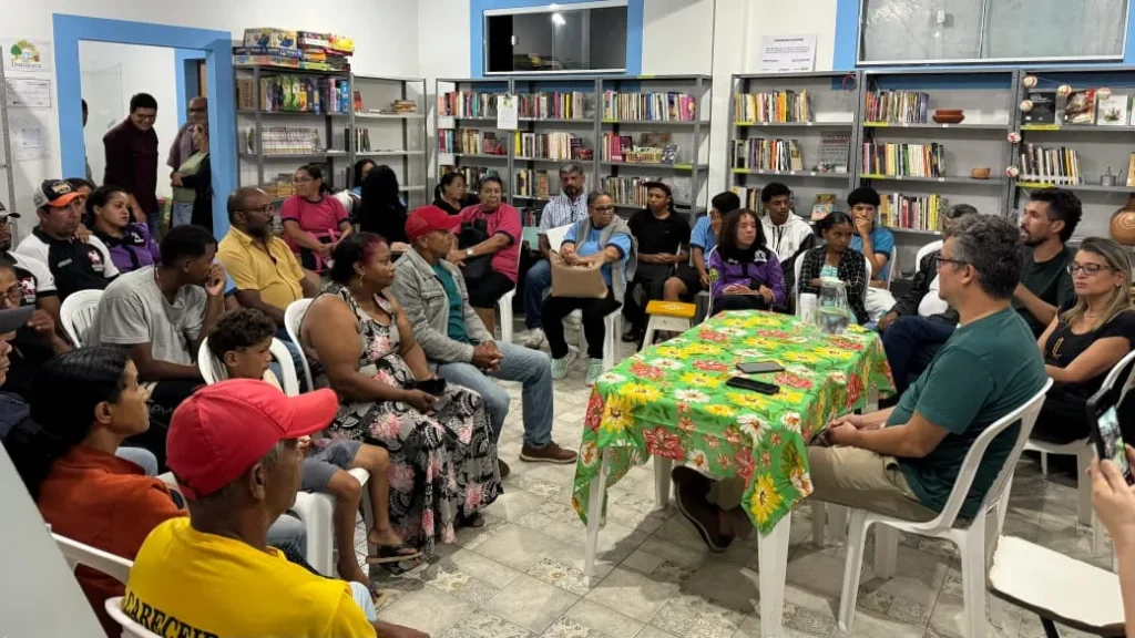 Dezenas de pessoas estão sentadas em cadeiras de plástico brancas dispostas em círculo durante uma reunião. No centro do espaço, há uma mesa retangular coberta por uma toalha verde estampada com grandes flores amarelas e vermelhas. 