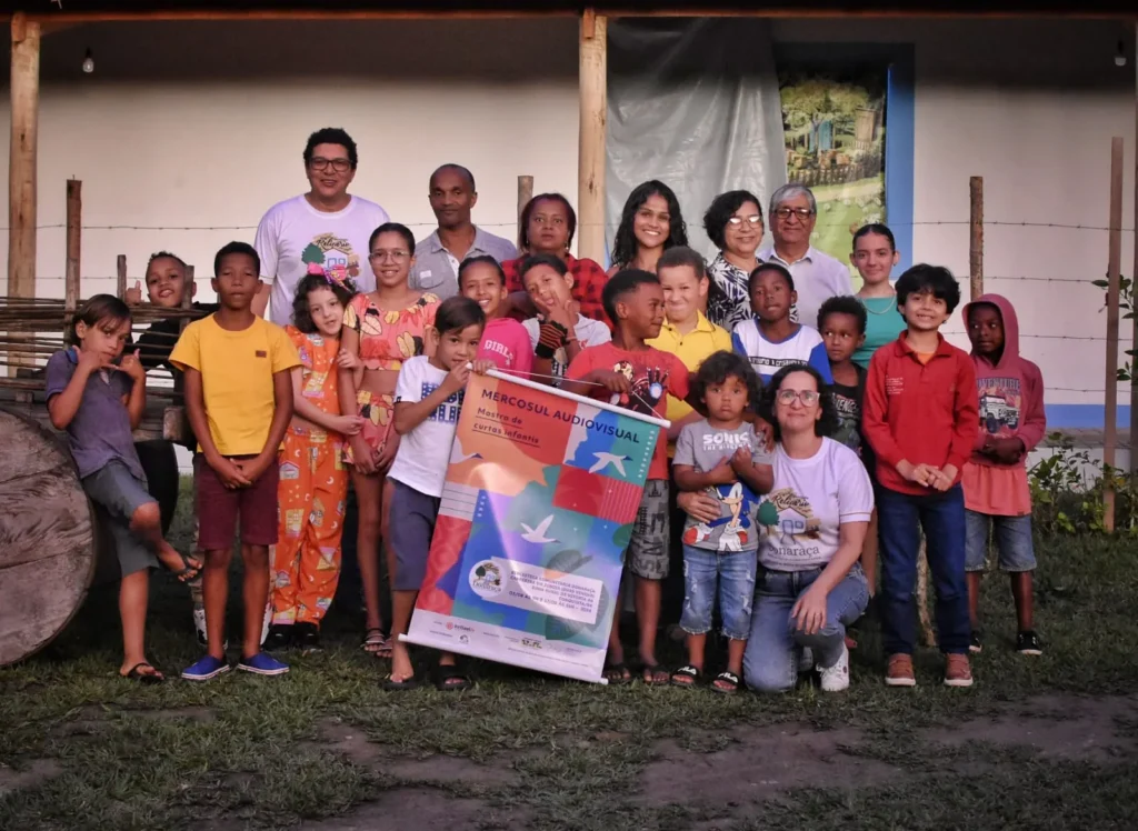 Grupo de crianças e adultos posa ao ar livre em frente a uma casa, reunido em torno de um banner colorido. As crianças ocupam a parte da frente e usam roupas de cores variadas, enquanto os adultos estão atrás, alguns vestindo camisetas com o logotipo do Instituto Relicário. No centro, o banner traz os textos “MERCOSUL AUDIOVISUAL” e “Mostra de curtas infantis”. 