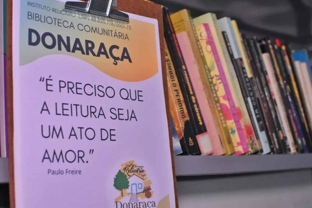 Um cartaz fixado em uma prancheta de madeira diz: É PRECISO QUE A LEITURA SEJA UM ATO DE AMOR, Paulo Freire. No canto inferior direito do papel, há a logomarca do Instituto Relicário Donaraça. Ao fundo, ligeiramente desfocada à direita, está uma prateleira cinza repleta de livros coloridos.