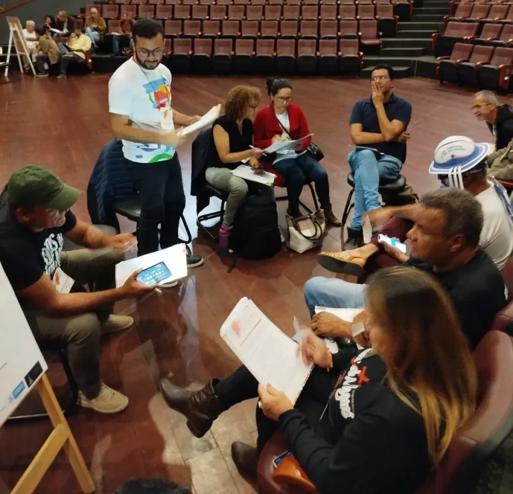 Grupo de pessoas reunidas em círculo em um auditório, participando de uma atividade de discussão. Algumas estão sentadas e outras em pé, segurando folhas e fazendo anotações. Ao fundo, há várias fileiras de poltronas vazias em tons de marrom e paredes revestidas de madeira. O ambiente é amplo, com piso de madeira escura e iluminação interna suave.