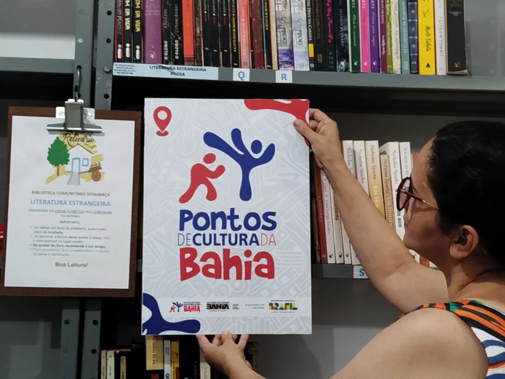 Mulher de óculos segura um cartaz diante de uma estante de livros. O cartaz tem fundo branco com elementos em vermelho e azul e traz o texto: “Pontos de Cultura da Bahia”. Na parte inferior aparecem os logotipos e os textos “Bahia”, “Governo da Bahia”, “Ministério da Cultura”, “Governo Federal Brasil” e “União e Reconstrução”. Ao lado, há uma placa presa à estante com o logotipo do Instituto Relicário e o texto “Biblioteca Comunitária Donaraça”, “Literatura Estrangeira” e “Boa Leitura!”. Ao fundo, livros organizados em prateleiras.