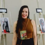Uma mulher sorridente com cabelos longos, escuros e ondulados posiciona-se no centro, entre dois cavaletes de madeira clara que expõem ilustrações em preto, branco e vermelho. Ela veste uma blusa de gola alta na cor marrom, calça preta e usa um crachá pendurado no pescoço. O fundo é uma parede lisa em tom claro. O crachá traz a identificação "Raquel Dantas" escrita à mão.