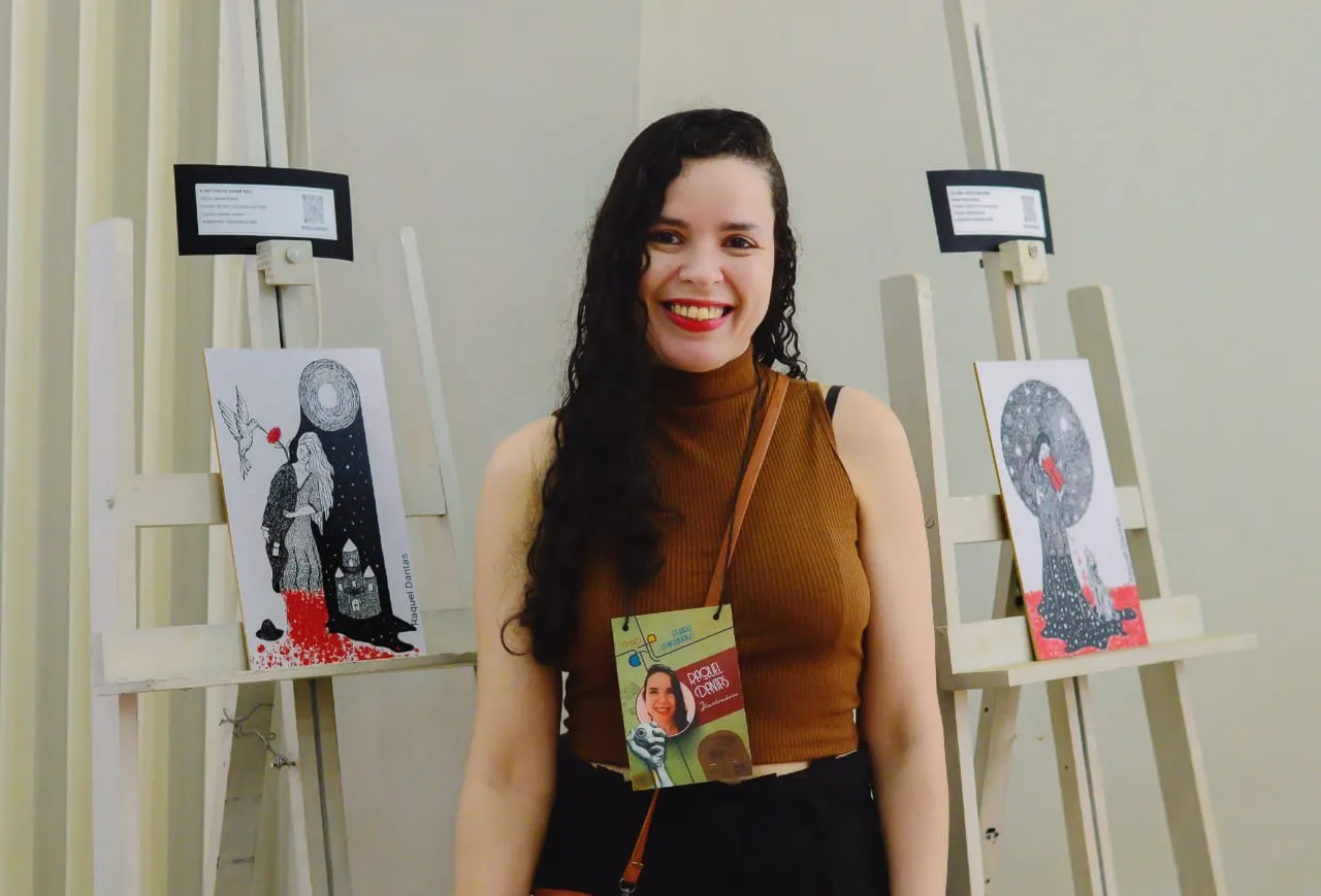 Uma mulher sorridente com cabelos longos, escuros e ondulados posiciona-se no centro, entre dois cavaletes de madeira clara que expõem ilustrações em preto, branco e vermelho. Ela veste uma blusa de gola alta na cor marrom, calça preta e usa um crachá pendurado no pescoço. O fundo é uma parede lisa em tom claro. O crachá traz a identificação "Raquel Dantas" escrita à mão.