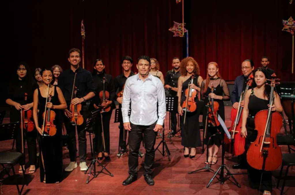 Um grupo de músicos de uma orquestra de cordas posa sorrindo em cima de um palco. No centro, um homem de camisa social branca e calça escura destaca-se à frente dos demais. Ao redor dele, cerca de doze músicos vestem roupas pretas e seguram instrumentos como violinos, violoncelos e um contrabaixo acústico.