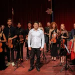 Um grupo de músicos de uma orquestra de cordas posa sorrindo em cima de um palco. No centro, um homem de camisa social branca e calça escura destaca-se à frente dos demais. Ao redor dele, cerca de doze músicos vestem roupas pretas e seguram instrumentos como violinos, violoncelos e um contrabaixo acústico.