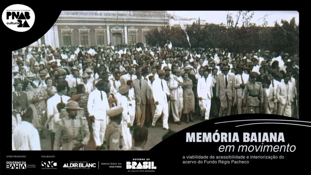 Fotografia em preto e branco de 1951. No canto esquerdo superior da imagem está a Prefeitura de Vitória da Conquista e o lado direito possui árvores. Régis Pacheco traja um terno branco, posicionado próximo ao centro da imagem e olha para o canto direito da foto. A imagem aparenta ter centenas de pessoas. Uma mulher de vestido longo está abraçada a Régis Pacheco. Militares fardados estão na ponta inferior esquerda e direita da foto, localizados próximos ao político, e criam um espaço mais vazio na parte direita inferior da fotografia. Instrumentos de sopro estão na multidão.Entre os trajes dos civis é possível identificar homens usando ternos, gravatas e chapéus, e mulheres que trajam vestidos lisos e estampados. Contornando toda a fotografia está posicionada uma margem preta com as informações: “Memória baiana em movimento, a viabilidade de acessibilidade e interiorização do acervo do Fundo Régis Pacheco”, em letra cinza, e as logos do Governo da Bahia, Sistema Nacional de Cultura, Política Nacional Aldir Blanc, Ministério da Cultura e Governo Federal.