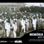 Fotografia em preto e branco de 1951. No canto esquerdo superior da imagem está a Prefeitura de Vitória da Conquista e o lado direito possui árvores. Régis Pacheco traja um terno branco, posicionado próximo ao centro da imagem e olha para o canto direito da foto. A imagem aparenta ter centenas de pessoas. Uma mulher de vestido longo está abraçada a Régis Pacheco. Militares fardados estão na ponta inferior esquerda e direita da foto, localizados próximos ao político, e criam um espaço mais vazio na parte direita inferior da fotografia. Instrumentos de sopro estão na multidão.Entre os trajes dos civis é possível identificar homens usando ternos, gravatas e chapéus, e mulheres que trajam vestidos lisos e estampados. Contornando toda a fotografia está posicionada uma margem preta com as informações: “Memória baiana em movimento, a viabilidade de acessibilidade e interiorização do acervo do Fundo Régis Pacheco”, em letra cinza, e as logos do Governo da Bahia, Sistema Nacional de Cultura, Política Nacional Aldir Blanc, Ministério da Cultura e Governo Federal.