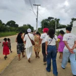 Um grupo de pessoas caminha de costas por uma calçada ao lado de uma rua pavimentada. Entre elas, algumas vestem camisas brancas. À esquerda, uma pessoa veste blusa branca e calça vermelha, acompanhada por duas crianças com vestidos vermelho e azul. No centro, sobressaem-se uma mulher com terno branco e turbante claro, outra com vestido estampado amarelo e floral, e uma pessoa de cabelo curto e rosa.