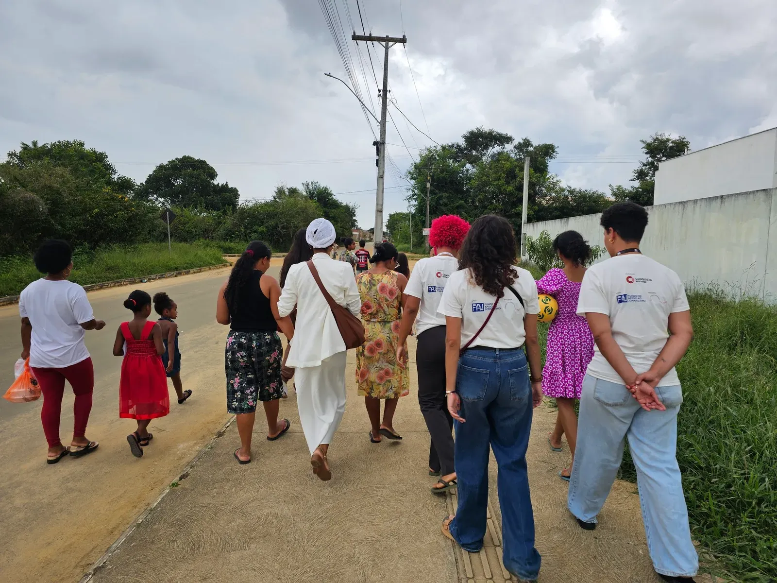 Um grupo de pessoas caminha de costas por uma calçada ao lado de uma rua pavimentada. Entre elas, algumas vestem camisas brancas. À esquerda, uma pessoa veste blusa branca e calça vermelha, acompanhada por duas crianças com vestidos vermelho e azul. No centro, sobressaem-se uma mulher com terno branco e turbante claro, outra com vestido estampado amarelo e floral, e uma pessoa de cabelo curto e rosa.