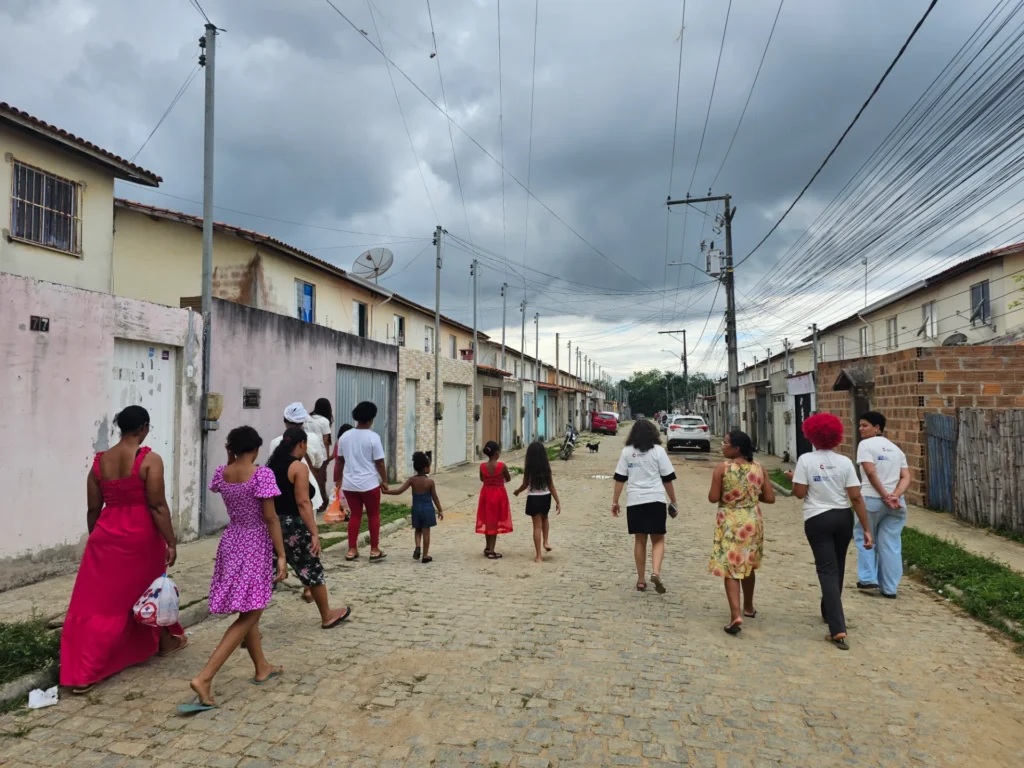 Um grupo de pessoas, composto majoritariamente por mulheres e crianças, caminha de costas por uma rua residencial pavimentada com paralelepípedos. Algumas pessoas vestem roupas coloridas em tons de rosa, vermelho e estampas florais, enquanto outras usam camisetas brancas. Dos dois lados da rua há casas simples, com muros e portões de diferentes cores. Fios elétricos cruzam o céu nublado e cinzento acima da via. Ao fundo da rua, carros estão estacionados.