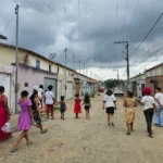 Um grupo de pessoas, composto majoritariamente por mulheres e crianças, caminha de costas por uma rua residencial pavimentada com paralelepípedos. Algumas pessoas vestem roupas coloridas em tons de rosa, vermelho e estampas florais, enquanto outras usam camisetas brancas. Dos dois lados da rua há casas simples, com muros e portões de diferentes cores. Fios elétricos cruzam o céu nublado e cinzento acima da via. Ao fundo da rua, carros estão estacionados.