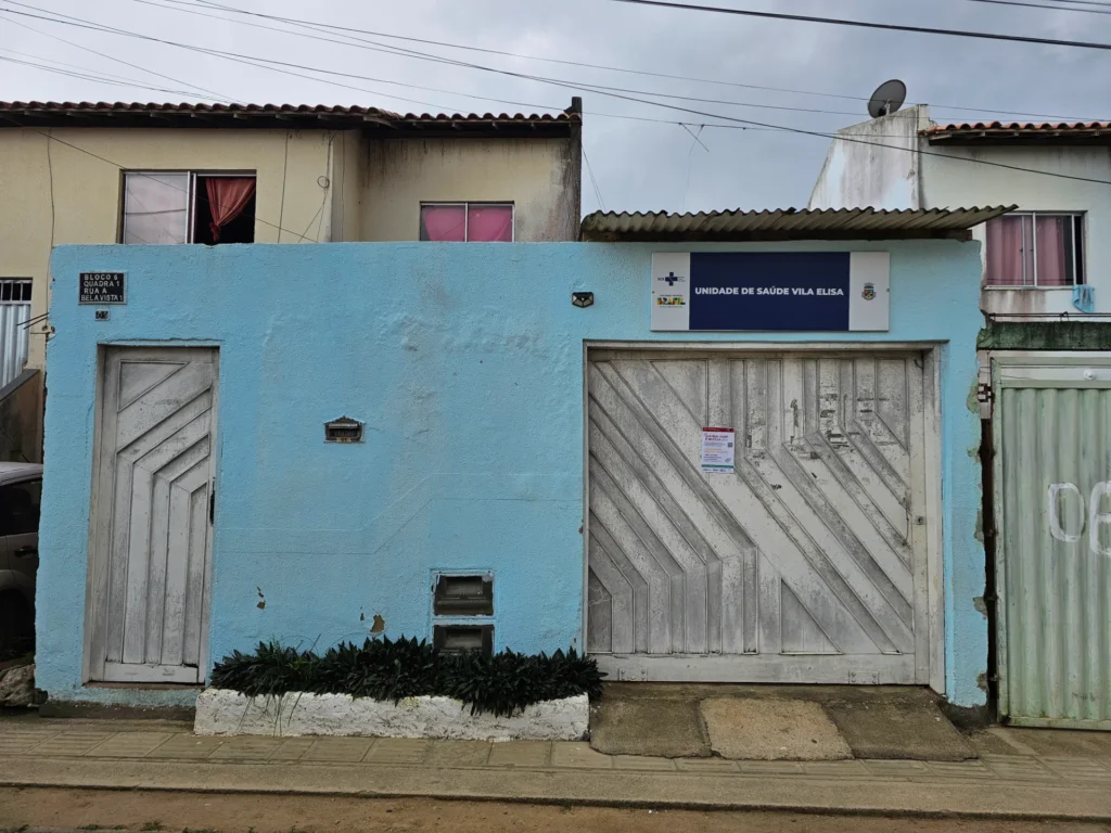 Frente térrea de um posto de saúde com muro azul claro e letreiro "UNIDADE DE SAÚDE VILA ELISA". Há um portão de ferro cinza à direita e uma porta menor à esquerda. Janelas com cortinas coloridas e telhados de casas vizinhas aparecem ao fundo sob céu nublado.