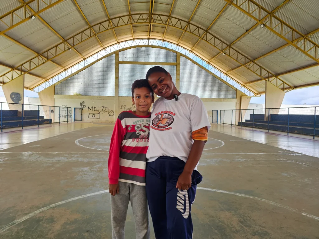 Uma mulher negra e um menino negro estão abraçados no centro de uma quadra de esportes coberta. A mulher, à direita, sorri com a cabeça inclinada, veste camiseta branca com estampa circular de capoeira e calça de moletom azul-escura. O menino, à esquerda, sorri, veste blusa de manga longa listrada em vermelho, cinza e branco, e calça de moletom cinza. 