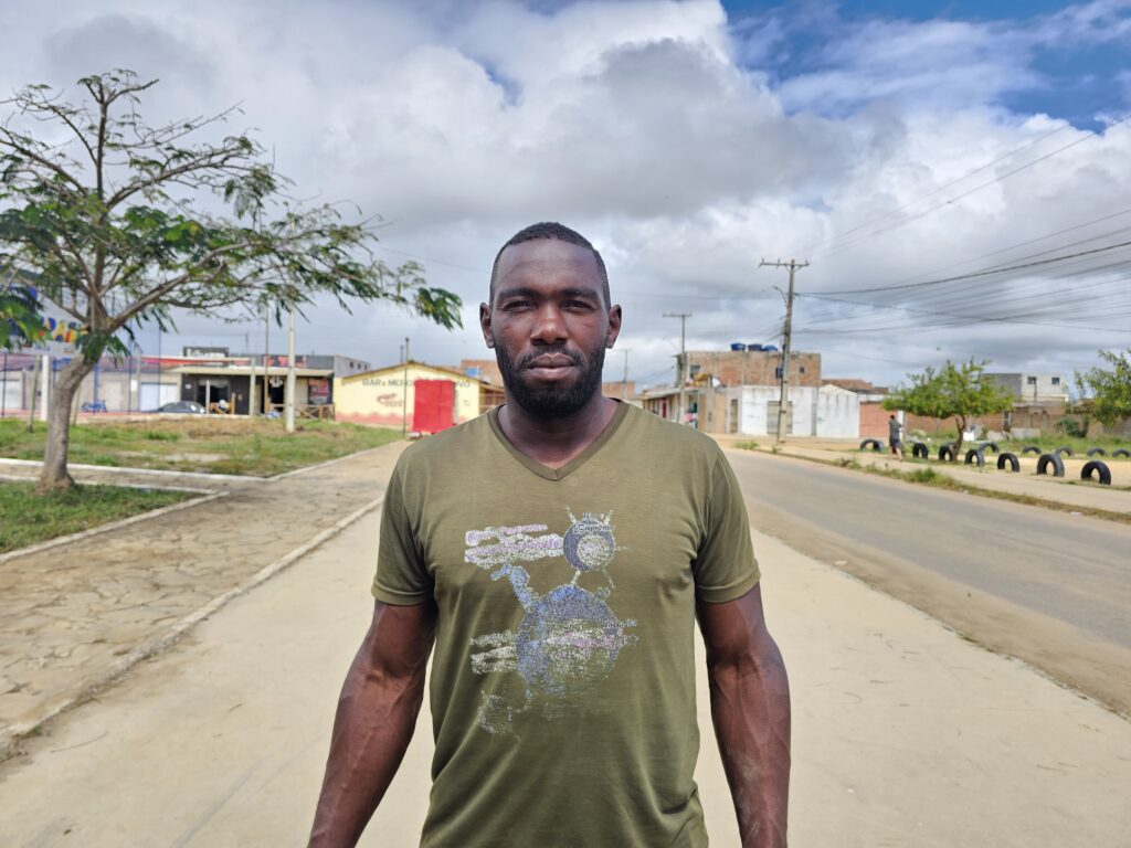 Um homem negro de barba curta e expressão séria está de pé em um calçadão. Ele veste uma camiseta verde. Ao fundo, uma estrada asfaltada segue pela direita e uma área de grama com árvores fica à esquerda. Mais atrás, há residências e pequenos comércios.