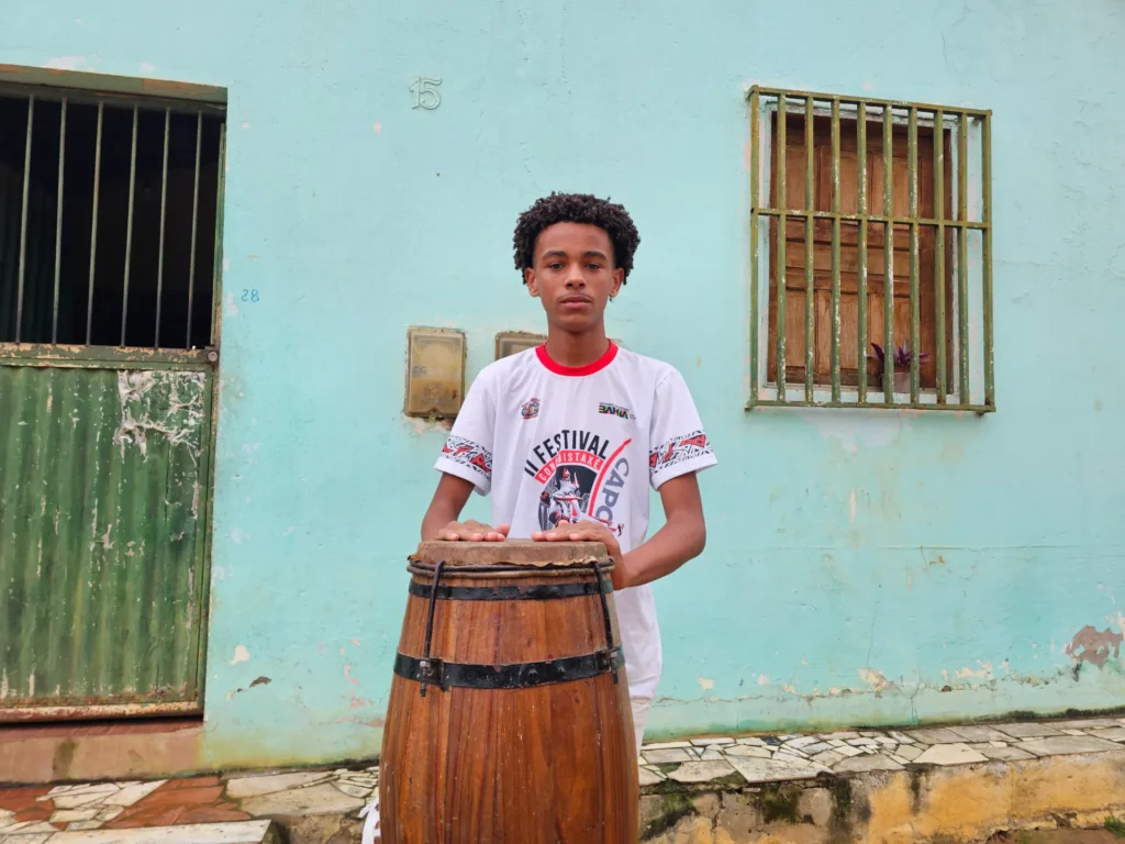 Um jovem negro de cabelo crespo curto está em pé, com as mãos apoiadas sobre um atabaque de madeira. Ele veste uma camiseta branca com detalhes em vermelho e preto, que traz a estampa "II FESTIVAL DE CAPOEIRA COQUINHO BAIANO" e a palavra "BAHIA". 