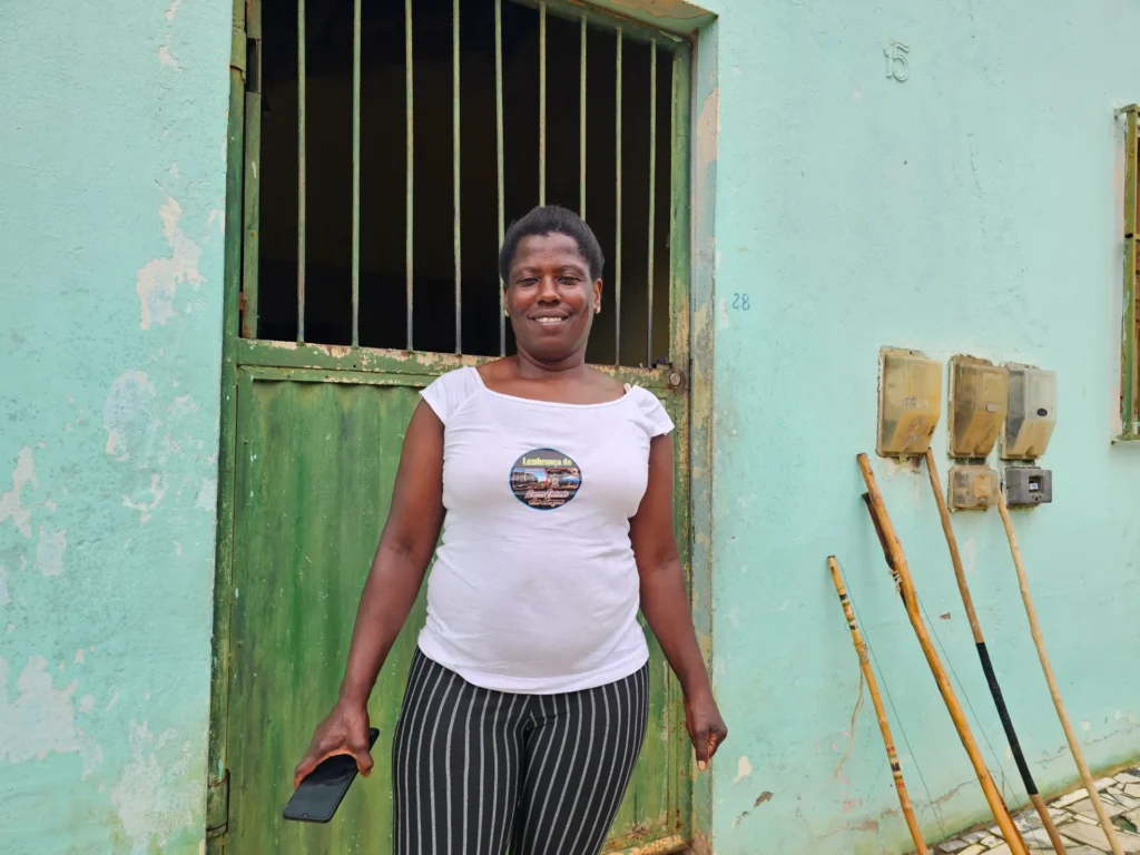 Uma mulher negra de cabelos curtos sorri em pé. Ela veste uma blusa branca de mangas curtas com uma estampa circular no peito e calça preta com listras verticais brancas. Na mão direita, ela segura um celular preto na altura da coxa. Ela está posicionada em frente a um portão de ferro verde com grades na parte superior.