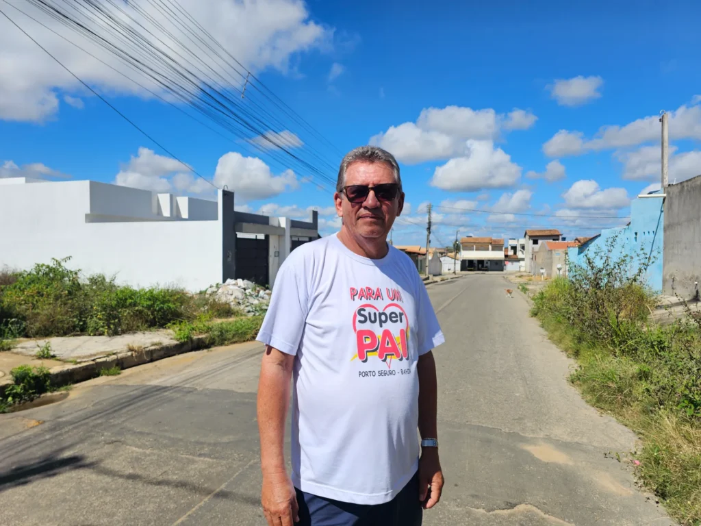 Um homem de óculos escuros está de pé no centro de uma rua residencial asfaltada. Ele veste uma camiseta branca com a estampa colorida "Para um Super Pai". Ao fundo, há casas simples sob um céu azul com nuvens brancas e fios elétricos.