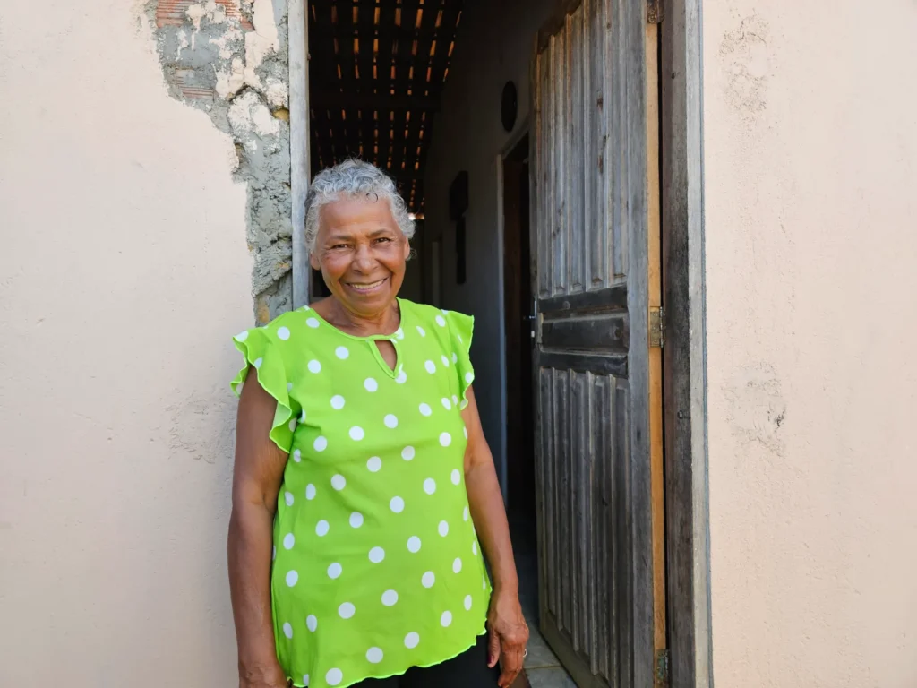 Uma mulher idosa e sorridente, de cabelos grisalhos curtos, posa na entrada de uma casa. Ela veste uma blusa verde-claro com estampa de bolinhas brancas. Atrás dela, destacam-se uma porta de madeira aberta e uma parede de tom bege.