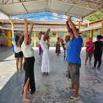 Pessoas de várias idades estão de mãos erguidas em um pátio coberto por uma estrutura de metal sustentada por vigas amarelas. Pequenas bandeirolas juninas decoram os arredores do espaço. O chão cimentado apresenta sinais de desgaste.