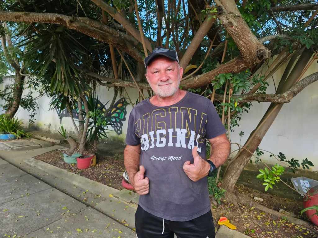 Um homem de meia-idade e barba branca sorri para a câmera, com os dois polegares para cima. Ele veste um boné e uma camiseta preta estampada. Ele está de pé em uma calçada com árvores e plantas ao fundo.
