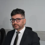 Vereador Diogo Azevedo veste terno preto, camisa social branca e gravata escura. Ele é um homem de pele clara com cabelos grisalhos, usa óculos de armação grossa e barba curta. Ele está sentado em uma cadeira de escritório.