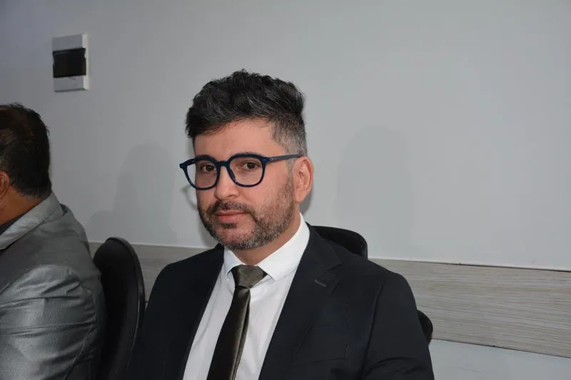 Vereador Diogo Azevedo veste terno preto, camisa social branca e gravata escura. Ele é um homem de pele clara com cabelos grisalhos, usa óculos de armação grossa e barba curta. Ele está sentado em uma cadeira de escritório.