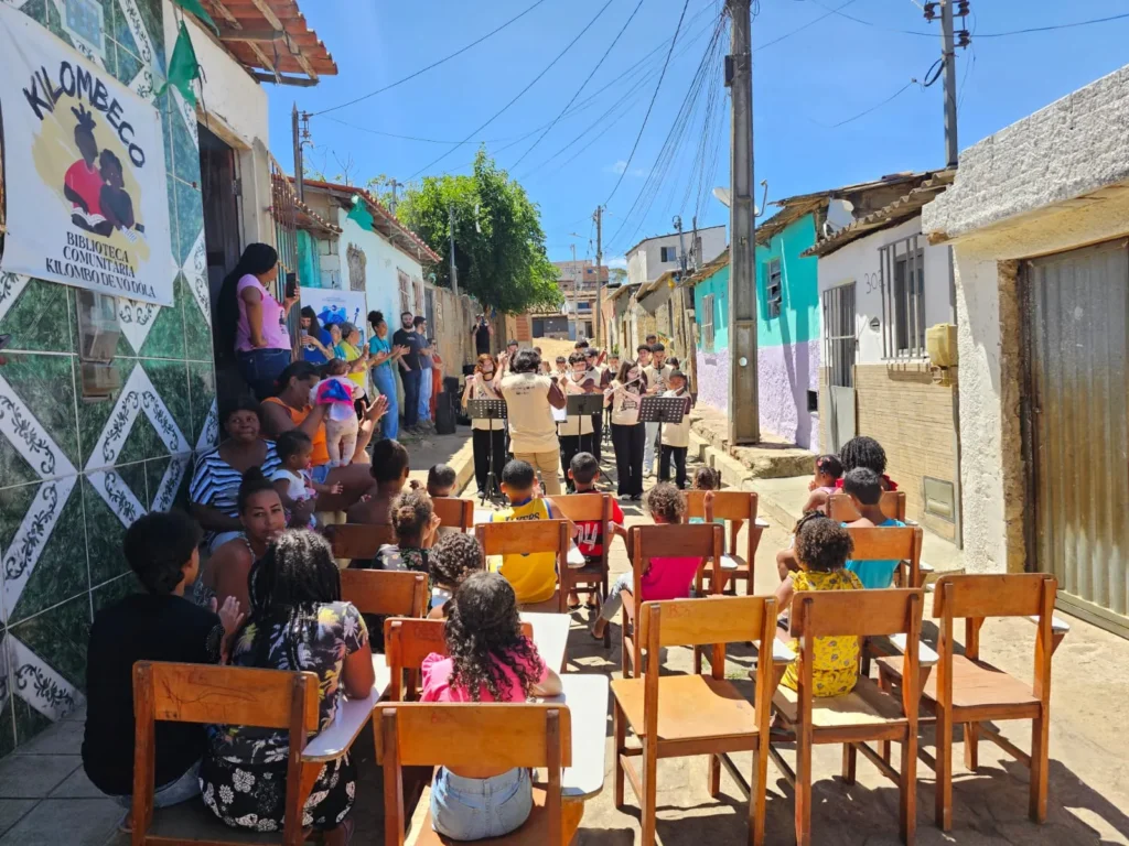 Crianças e adultos assistem a uma apresentação musical em uma rua estreita sob o céu azul. A plateia está sentada de costas em cadeiras escolares de madeira organizadas no meio da via. Ao fundo, um grupo de jovens em pé toca instrumentos de sopro, como flautas e clarinetes, posicionados atrás de estantes de partituras pretas.