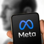 Uma mão segura um smartphone. A tela do celular exibe o logotipo azul da Meta e a palavra "Meta" em branco sobre fundo preto. Ao fundo, desfocado, vê-se o perfil de Mark Zuckerberg.