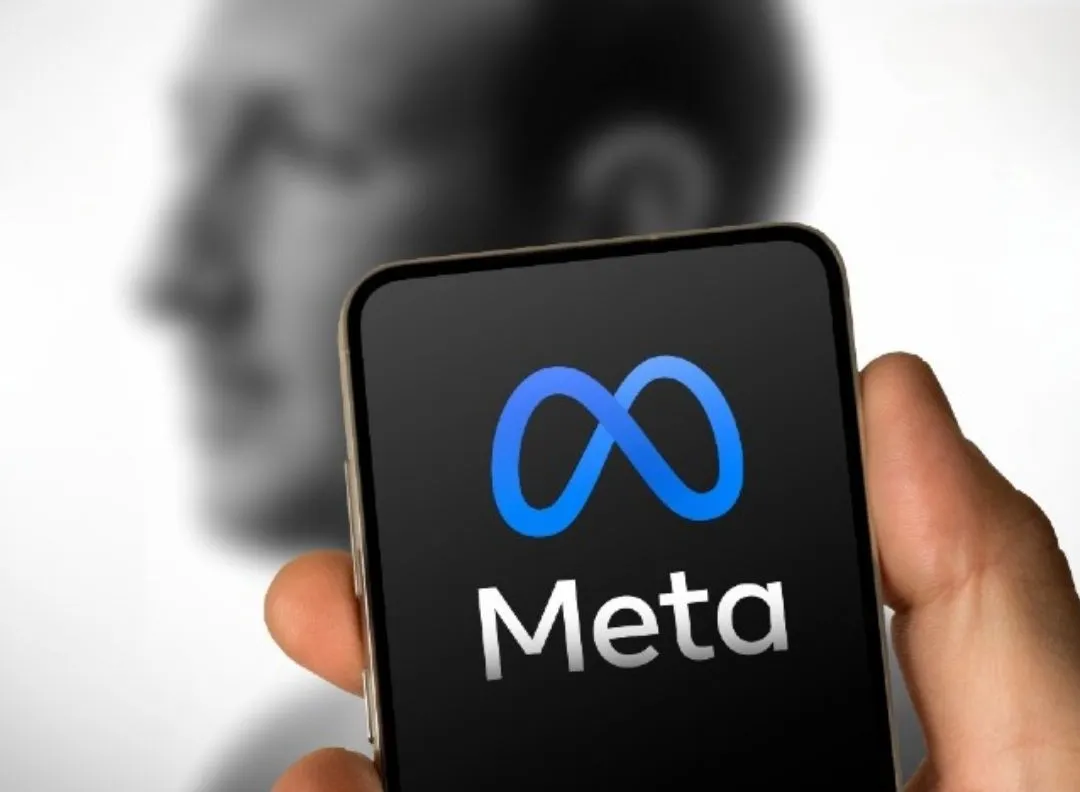 Uma mão segura um smartphone. A tela do celular exibe o logotipo azul da Meta e a palavra "Meta" em branco sobre fundo preto. Ao fundo, desfocado, vê-se o perfil de Mark Zuckerberg.