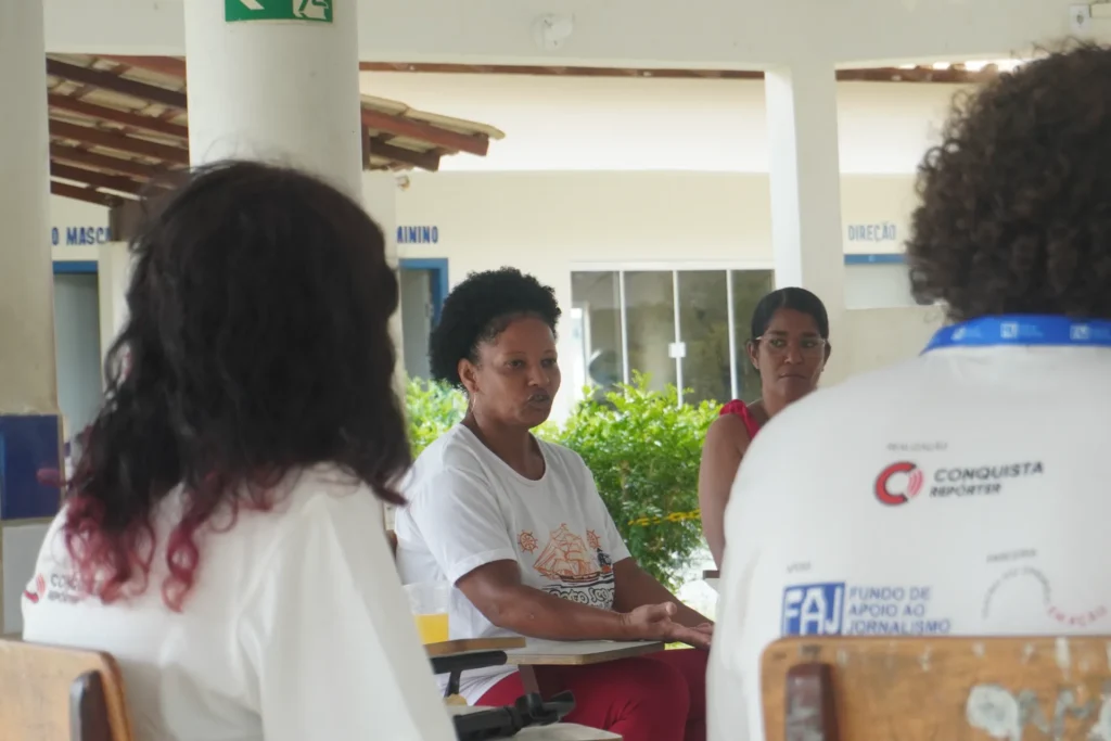 Uma mulher de camiseta branca com estampa de barco está sentada e falando em um ambiente aberto. Ela é ladeada por duas pessoas de costas, que vestem camisetas brancas com logotipos do 'Conquista Repórter' e 'FAJ'. Uma quarta pessoa está visível ao fundo, atrás dela.