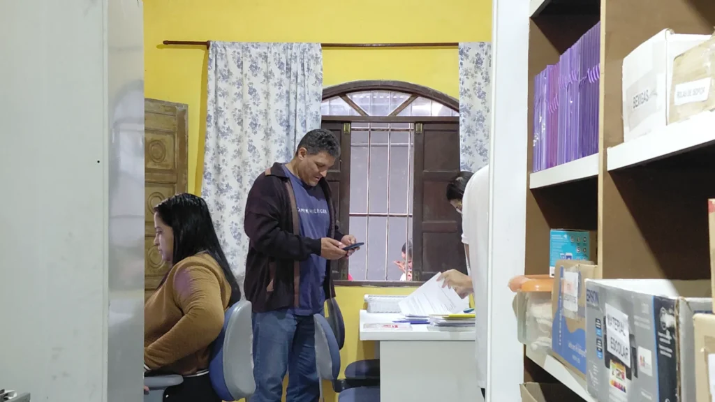 Fotografia colorida horizontal capturada em plano médio-longo mostra três pessoas em um escritório simples com paredes amarelas. Ao centro, um homem de casaco escuro em pé olha para seu celular, uma mulher de jaqueta marrom está sentada à sua esquerda e outra mulher em pé, de branco, está manuseando papéis sobre uma mesa à direita. Ao fundo, uma janela de madeira com grades e, à direita, estantes com caixas e arquivos organizados.