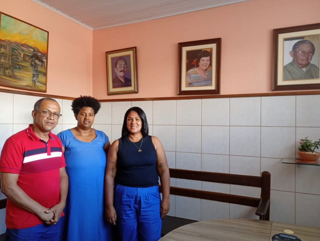 Três pessoas estão em pé, lado a lado, em uma sala com paredes salmão e azulejos brancos. À esquerda, um homem negro de óculos veste camisa polo vermelha com listras horizontais escuras e calça azul. No centro, uma mulher negra com cabelo crespo curto veste um vestido longo azul. À direita, uma mulher de pele morena e cabelos pretos lisos veste blusa regata preta e calça azul. Atrás deles, há um banco de madeira escura encostado na parede. Na parte superior da parede salmão, estão pendurados um quadro com uma pintura de paisagem rural e três retratos emoldurados com fotos de rostos de pessoas mais velhas.