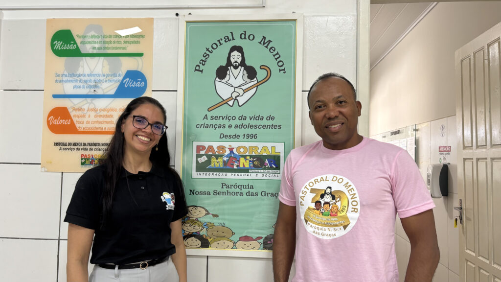 Uma mulher de pele clara e um homem negro. sorriem lado a lado em frente a uma parede branca com cartazes. À esquerda, a mulher usa óculos de grau, camisa polo preta e calça cinza claro. À direita, o homem veste uma camiseta rosa claro com uma estampa circular amarela que traz o desenho de Jesus com crianças e o texto "Pastoral do Menor - 30 Anos - A Serviço da Vida de Crianças e Adolescentes - Paróquia N. Sra. das Graças".
